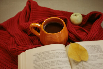 Obraz premium An orange mug of coffee resting on a red knitted sweater beside an open book and a yellow autumn leaf, with soft film grain, high quality photo
