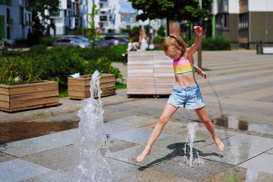 A joyful young girl jumping over water jets from a ground fountain in a city square on a warm sunny day