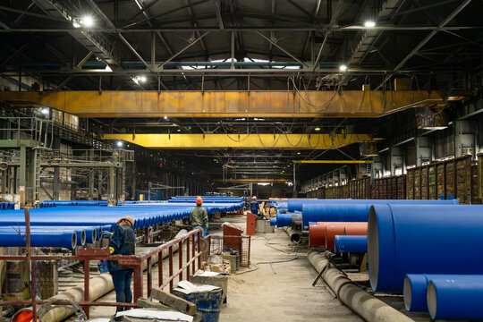 Ductile iron pipe manufacturing factory with workers among coated waterworks pipes, overhead cranes and heavy industrial production equipment.