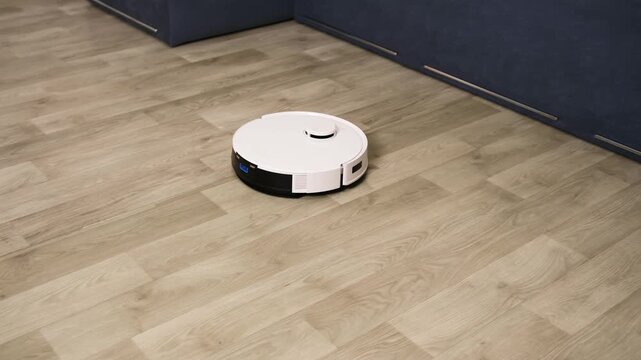 Modern white robotic vacuum cleaner autonomously moving on a laminate floor, efficiently cleaning the surface along the furniture. Smart home technology simplifying household chores for a tidy home
