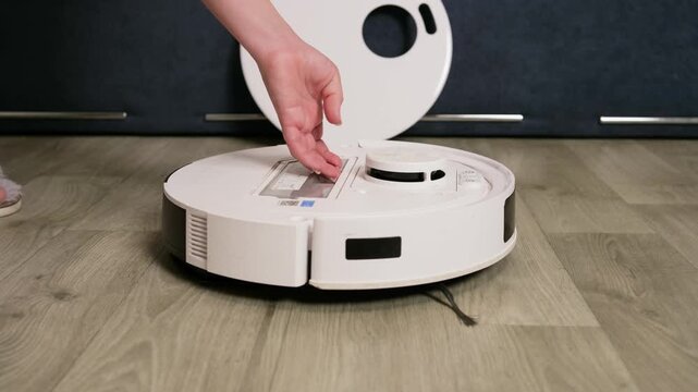 Female hands opening a modern robotic vacuum cleaner to remove and clean the dustbin container and filter, performing regular maintenance for a smart home appliance on a wooden floor