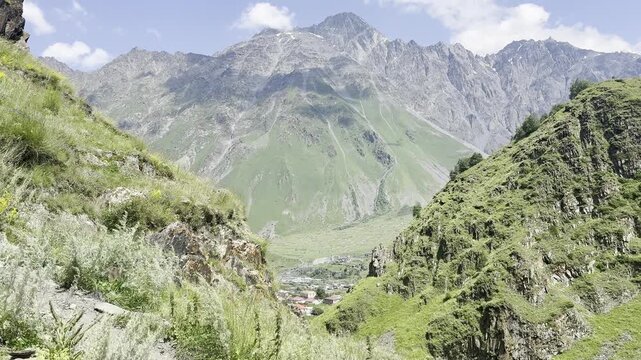 Scenic view of a mountain hiking trail overlooking a small village in a green valley with majestic peaks in the background under a blue sky
