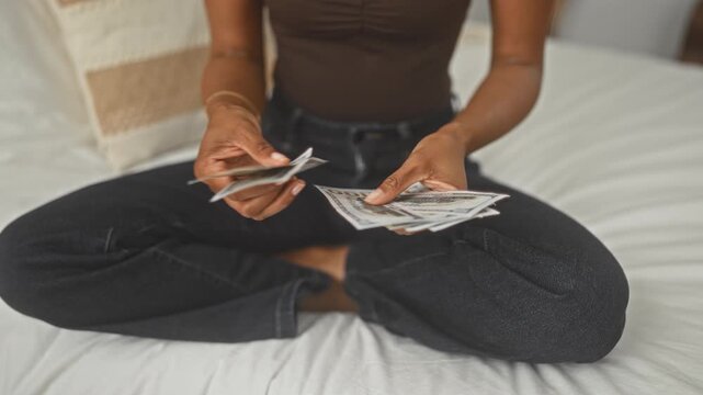 Young woman counting dollars with hands visible seated crosslegged on bed in a building; financial confidence.