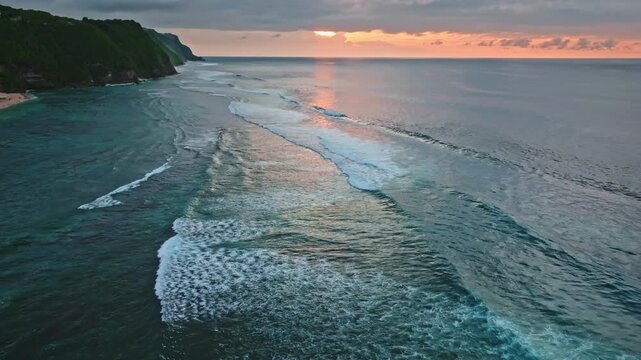 Bukit Peninsula coastline with green cliffs, sandy beach and clear turquoise ocean water with waves breaking, reflecting the golden light of tropical sunset. Travel and relax on Bali Island, Indonesia