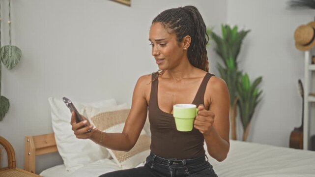 Woman holding smartphone and green mug while seated on bed in bedroom, glancing at screen with startled expression; surprise morning routine.