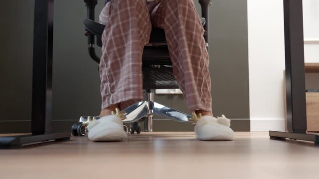 Person wearing plaid pajamas and plush reindeer slippers walks to office chair and sits down at desk. Concept of comfortable clothing and working from home