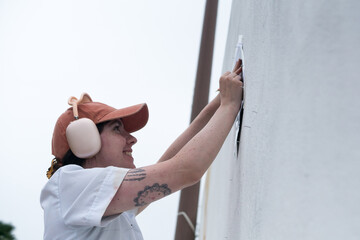 Smiling profile of a young female mural artist with an arm tattoo, cap, and large pink headphones, working with her hands on a large outdoor wall stencil for street art. © Phoscar