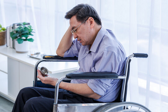 A middle-aged Asian man, a patient, sits alone in a wheelchair, holding tablets, pills, medications drugs, next to the window of his hospital room. There is space for a message.