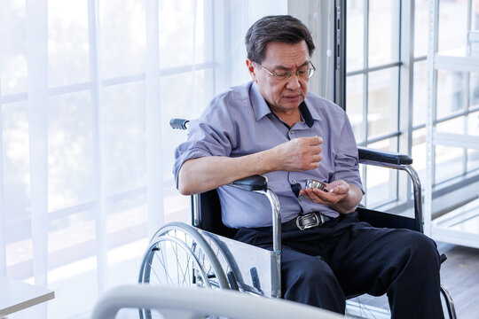 A middle-aged Asian man, a patient, sits alone in a wheelchair, holding tablets, pills, medications drugs, next to the window of his hospital room. There is space for a message.