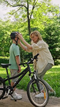 Blonde mother checks helmet on boy near bicycle on park path