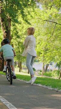 Mother claps as young boy rides bicycle on sunny park path