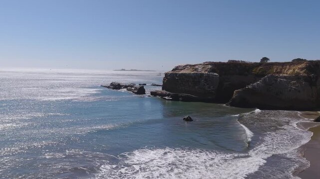 Aerial perspective reveals the rugged Pacific Ocean coastline by Franklin Point in California, where gentle waves roll onto a sandy beach beside rocky bluffs and reefs.