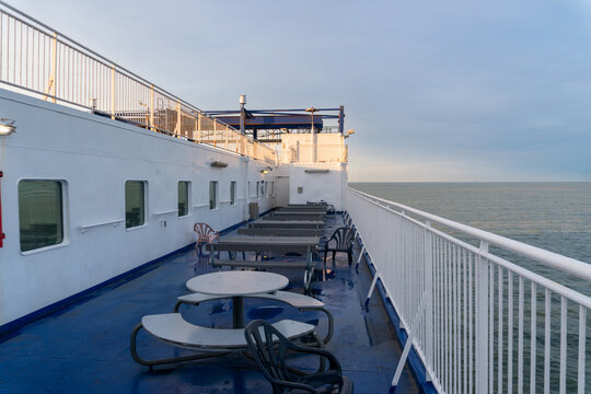 Quiet exterior promenade deck of a passenger ferry with picnic tables, chairs, white railings and panoramic sea view. Wet flooring and soft evening light create a peaceful onboard environment for mari
