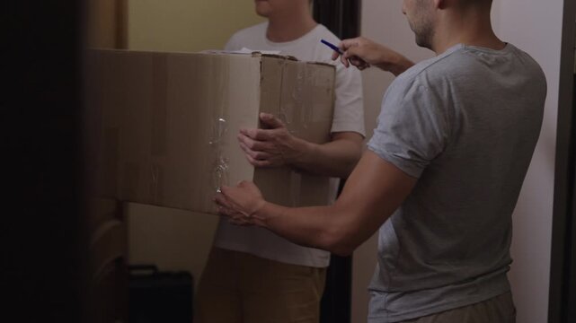 Courier delivering big parcel to man at apartment door. Customer takes box and signs delivery confirmation papers. Home delivery service concept. Shooting in slow motion.