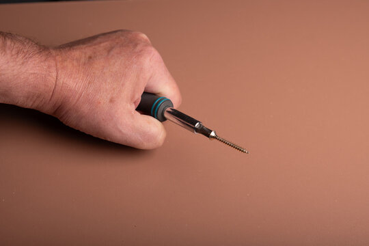 A human fist is holding a screwdriver and a screw is magnetic connectet to the phillips screwhead. A wooden board as background with space for text or digits.