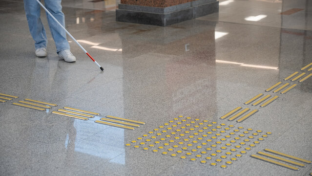 A blind woman walks on tactile paving. Close-up of her feet and a walking stick.