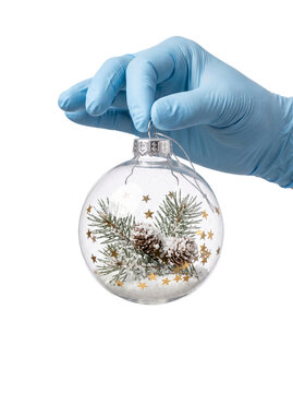 A hand wearing a medical glove holding a transparent background glass Christmas ornament with pine branches and snow inside