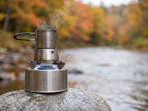 metal coffee pot heating up on a burner stove on a stone in the middle of a mountain river in autumn forest with hot steam