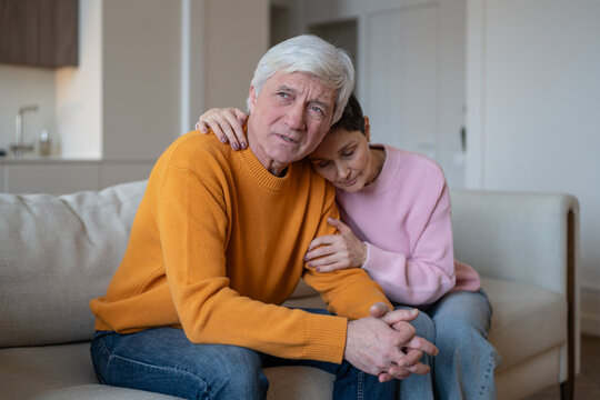 Senior couple sitting on sofa at home, loving wife leans against thoughtful husband and embraces partner, elderly spouses share closeness, emotional comfort and support during difficult life worries.