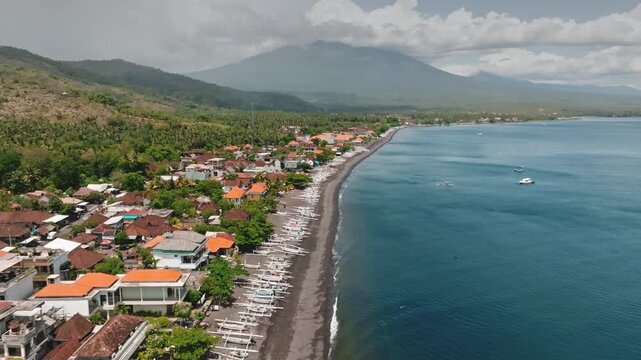 Amed beach, Bali coastline with traditional jukung fishing boats on black sand, village hugged by tropical greenery and dramatic Mount Agung volcano under cloudy blue sky. Aerial zoom out drone flight