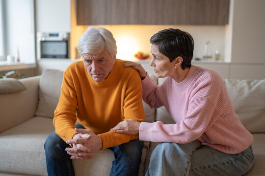 Senior couple sitting on sofa at home, caring wife touches shoulder of distressed husband and holds forearm, worried woman comforts upset partner, emotional support, empathy and family unity.