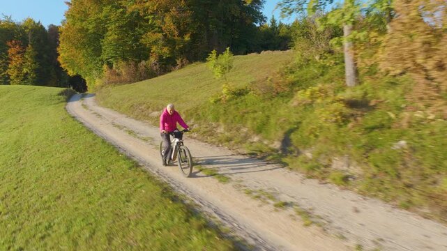 AERIAL Active elderly woman cycles toward camera on electric bike along sunlit gravel road leading through rolling grassland and woodland with autumn leaves. Outdoor exercise and healthy senior living