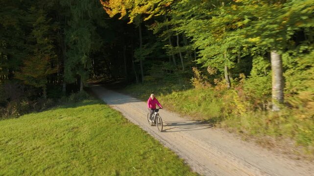 AERIAL: Fit senior woman rides an ebike from a shaded forest onto a sunny gravel road beside a green meadow. Elderly lady biking through peaceful autumn countryside among colourful turning leaves.