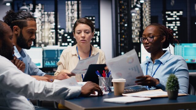 Diverse employee team reviews paperwork and company documents in the office late at night, analyzing budgets and documentation files while cooperating on reports before a business deadline.