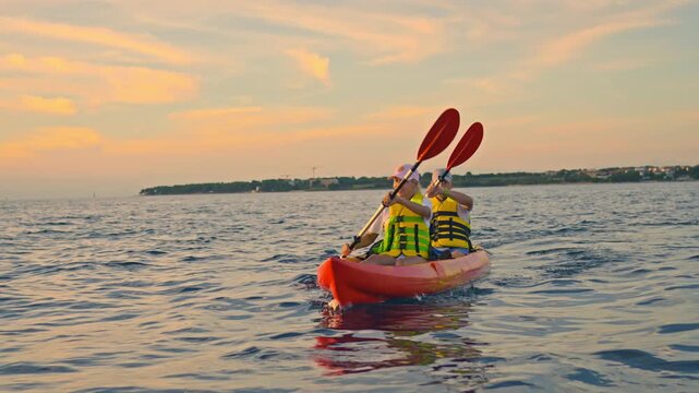 A couple enjoys a serene sea kayaking adventure during a picturesque sunset, paddling through calm waters in vibrant life vests and embracing nature's beauty.
