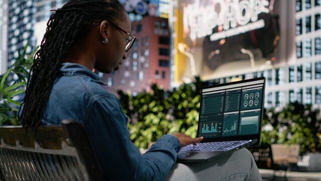 Black woman analyzes investment statistics and charts on a laptop while sitting on a park bench, working remotely outdoors and reading financial data from a dashboard interface to solve tasks.