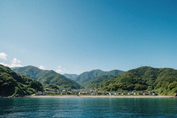 Fototapeta premium Aerial view of coastal village near blue ocean. Green mountains surround small houses by seaside shore. Calm sea water meets rocky coast under clear blue sky. Scenic summer landscape shows peaceful