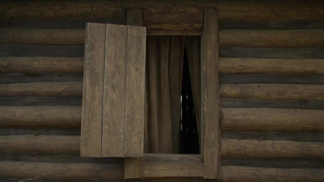 Wooden shutters and a burlap curtain cover the window of a log cabin. Historical rural architecture, traditional lifestyle, and off-grid dwelling.
