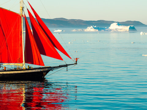 Greenland - 06 July 2026: Aerial view of a sailboat with striking red sails navigating through icy waters with icebergs in the background and a person standing on the bowsprit.