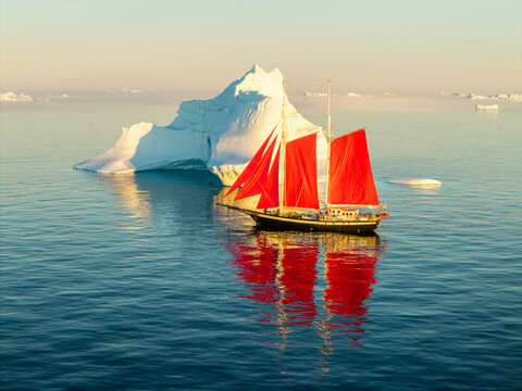 Aerial view of a sailboat with striking red sails navigating calm arctic waters near a massive white iceberg in Greenland.