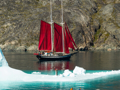 Aerial view of a sailboat with striking red sails navigating near a large iceberg in the icy waters of Greenland.