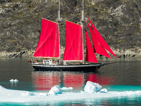 Aerial view of a schooner with vibrant red sails navigating through icy waters near a rocky coastline in Greenland.