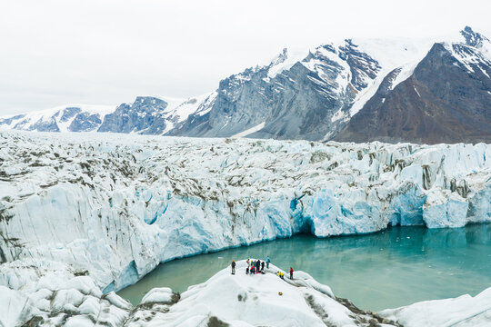 Aerial view of a massive glacier with deep blue ice formations and people standing on a rocky outcrop near a turquoise glacial lake, surrounded by snow-capped mountains in Greenland.