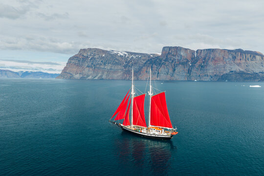 Aerial view of a schooner with striking red sails sailing through the deep blue waters near the dramatic rocky cliffs of Uummannaq, Kitaa, Greenland.