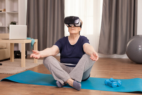 Senior woman uses a VR headset while seated on a yoga mat beside dumbbells, blending fitness technology, home exercise and healthy aging.
