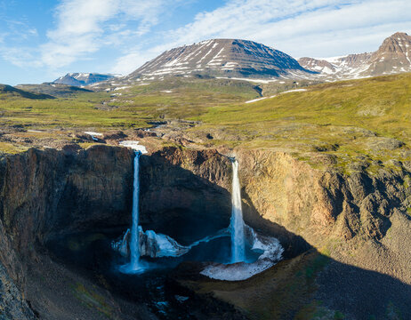 Aerial view of two waterfalls cascading into a pool with snow and ice, surrounded by rocky terrain and mountains in Greenland.