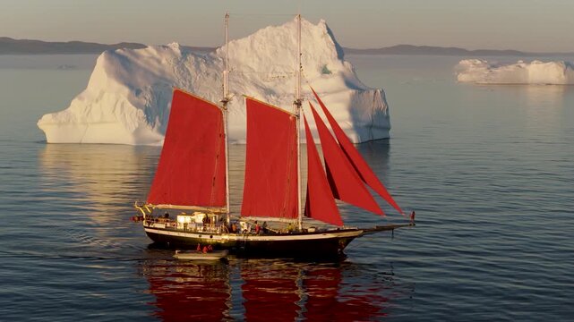 Aerial view of a schooner with red sails sailing past a massive iceberg in the calm arctic waters of Greenland.
