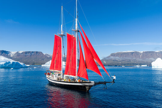 Aerial view of a sailboat with striking red sails navigating through Arctic waters surrounded by massive icebergs and rugged mountains in Greenland.