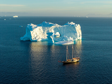 Aerial view of a massive arch iceberg floating in the deep blue waters of the Arctic with a sailboat nearby under a clear sky in Greenland.