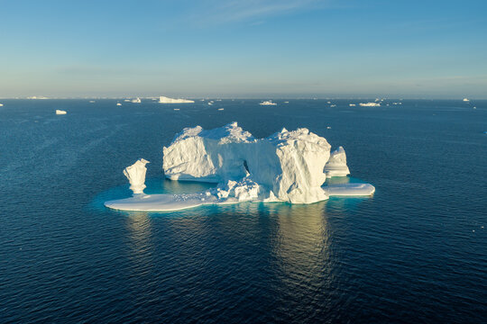 Aerial view of a massive arch iceberg floating in the deep blue waters of the Arctic Ocean, surrounded by smaller ice fragments under a clear sky in Greenland.
