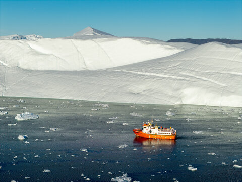 Aerial view of Ilulissat glacier with a small orange boat navigating through icy waters under a clear blue sky in Greenland.