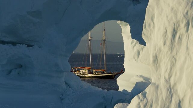 Aerial view of a sailboat framed by a natural arch in a massive iceberg floating in the cold waters of Greenland.