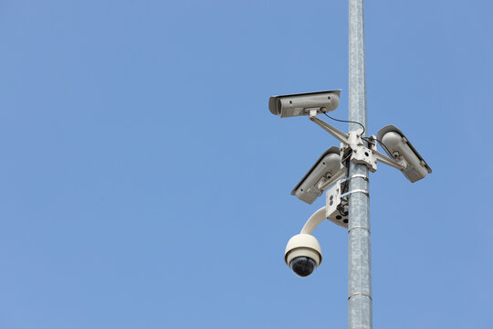 Security cameras and surveillance equipment mounted on tall metal pole against clear blue sky, featuring multiple fixed lenses and dome camera in urban infrastructure