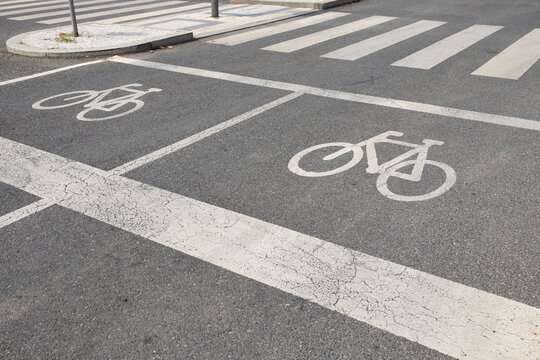 Bicycle lane markings and zebra crossing on urban asphalt street with painted cycling symbols, road lines, sidewalk island, and traffic infrastructure in city roadway