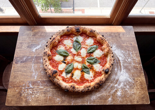 Pizza margarita napolitana reci&eacute;n horneada sobre mesa de madera con harina junto a la ventana