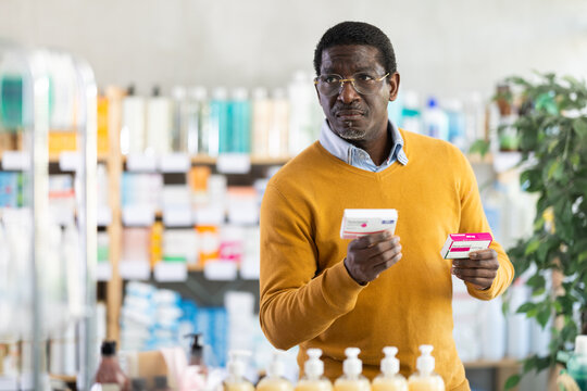 African man choose buy painkiller paracetamol tablet at pharmacy, compare with other good. Client check for absence of harmful substances, view product in details.